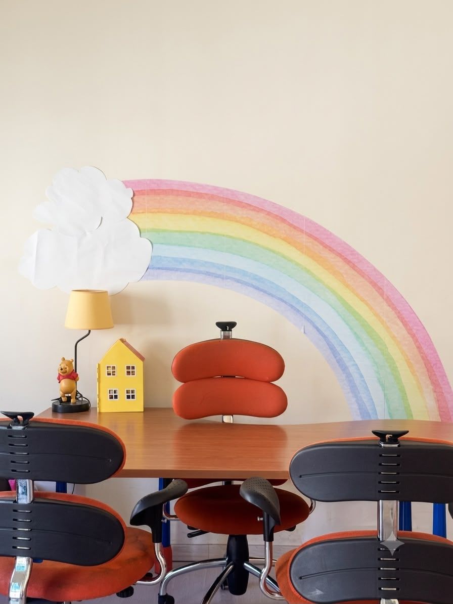 Tavolo con sedie colorate per le attività, davanti a un murale con un arcobaleno, una lampada di Winnie the Pooh e una casetta gialla